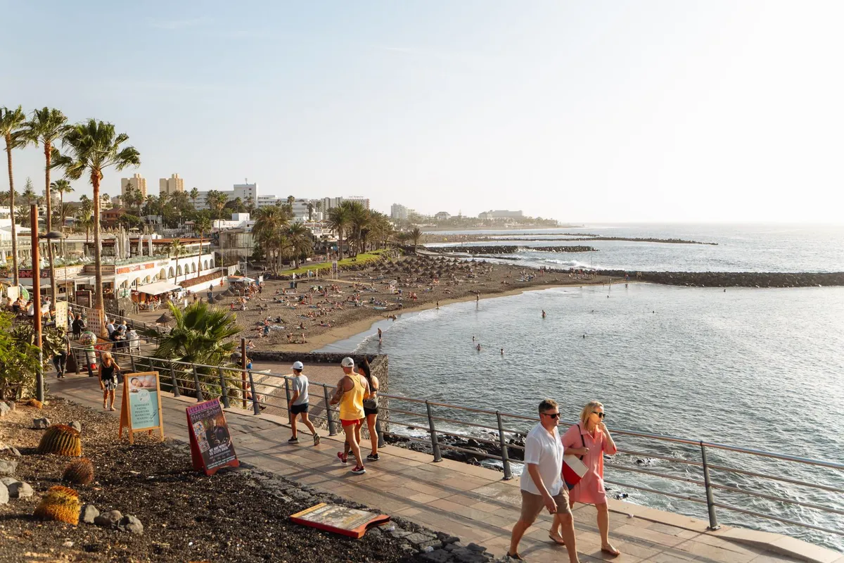 Tourists walking the seafront promenade past a resort beach on Tenerife's south coast in afternoon sun.